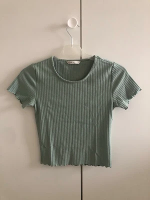 Ribbad grön croppad topp från ONLY - Snygg ribbad croppad topp från ONLY i ljusgrön färg. Toppen har korta ärmar, rund halsringning och vågig kant nertill och vid ärmslut. Perfekt att matcha med höga jeans eller kjol.
