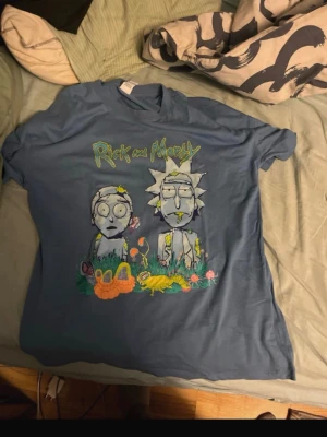 Blå Rick and Morty t-shirt - Cool blå t-shirt med Rick and Morty-motiv från Adult Swim. Trycket visar Rick och Morty bland färgglada växter och svampar. T-shirten har korta ärmar och är gjord i mjuk bomull, perfekt för fans av serien.