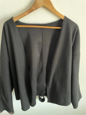 Svart kappa med knytband - Minimalistisk svart kappa med öppen front och vida ärmar. Kappan har ett brett knytband. Tillverkad i ett mjukt tyg som känns bekvämt och har en rak, avslappnad passform. Perfekt för lager på lager och enkel att styla.