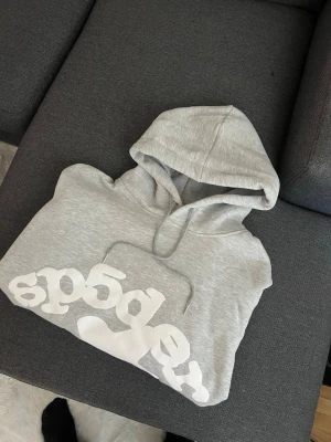 Grå hoodie från SP5DER med tryck - Helt oanvänd Snygg ljusgrå hoodie från SP5DER med stor vit logga i coolt tryck framtill. Klassisk modell med huva och dragsko, tillverkad i mjukt material som känns skönt mot huden. Perfekt för en avslappnad streetwear-look.