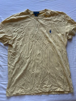 Gul t-shirt från Ralph Lauren - Snygg gul t-shirt från Ralph Lauren med klassisk blå logga broderad på bröstet. T-shirten har rund halsringning och korta ärmar