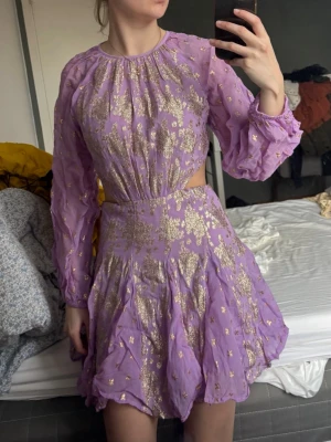 Lila och guldglittrig kortklänning - Superfin lila kortklänning med glittriga gulddetaljer och långärmade ärmar. Klänningen har en öppen detalj i midjan, öppen rygg och volangig kjol. 