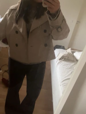  beige trenchcoat - Snygg kort trenchcoat i beige med dubbelknäppning och stora svarta knappar. Jackan har klassisk krage, bälte med spänne vid ärmsluten och en rak, boxig passform. Perfekt för dig som gillar stilrena och tidlösa plagg.