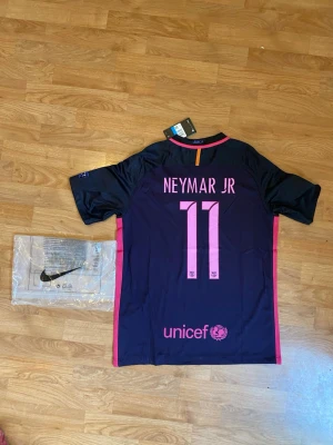 Barcelona Neymar Jr matchtröja Nike 2016/17 - Snygg Barcelona matchtröja från Nike med Neymar Jr och nummer 11 på ryggen. Tröjan är nytt, mörkblå med rosa detaljer, klubbmärke och Qatar Airways-tryck på bröstet samt Champions League-märke på ärmen. Tillverkad i lätt och ventilerande material. 