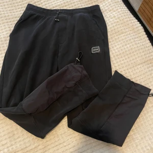 Svarta mjukisbyxor från Hugo - Svarta mjukisbyxor från Hugo med elastisk midja och dragsko. Byxorna har ribbade muddar vid bensluten och en diskret logotyp på vänster ben. Tillverkade i mjukt material som känns skönt mot huden. Perfekta för en avslappnad stil.