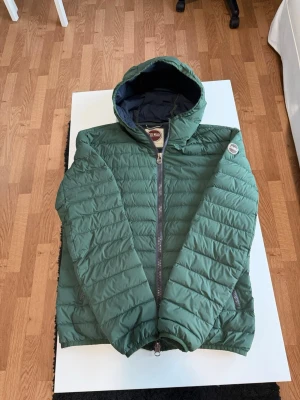 Colmar jacka - Snygg grön pufferjacka från Colmar med huva och svarta detaljer. Jackan har quiltad design, tvåvägsdragkedja och två fickor med dragkedja. Perfekt för kyliga dagar och ger en sportig look. Märke på ärmen och fodrad insida för extra värme.