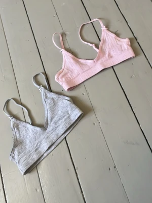 Två pack toppar från H&M - Två bekväma braletter från H&M i storlek 134/140. En i ljusgrått och en i ljusrosa, båda med smala axelband och mjuk, stretchig bomullsblandning. Perfekta för en enkel och avslappnad stil.