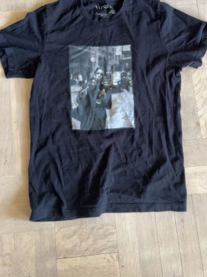 Svart t-shirt med tryck från Vonix - Svart t-shirt från Vonix med ett stort tryck framtill som föreställer en person i stadsmiljö. Klassisk passform med rund halsringning och korta ärmar. Perfekt för dig som gillar streetstyle och grafiska motiv.