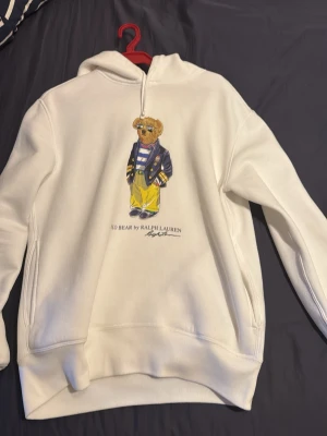 Vit hoodie Polo Bear Ralph Lauren - Vit hoodie från Ralph Lauren med ikoniska Polo Bear-trycket på bröstet. Klassisk modell med huva och känguruficka. Mjuk bomullskvalitet och avslappnad passform, perfekt för en chill och stilren look.