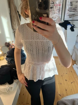 Vit spetsblus med volang - Superfin vit spetsblus med korta ärmar och volang nertill. Blusen har rund halsringning och ett vackert spetsmönster över hela plagget. Perfekt att styla med jeans eller kjol för en trendig look.