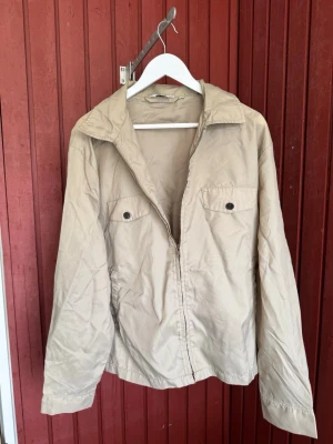 Beige jacka från Levi's i storlek XL - Snygg beige jacka från Levi's med klassisk krage och två bröstfickor med knappar. Jackan har dragkedja framtill och är tillverkad i ett lätt material som passar perfekt till vår och höst. Enkel och stilren design som funkar till det mesta.