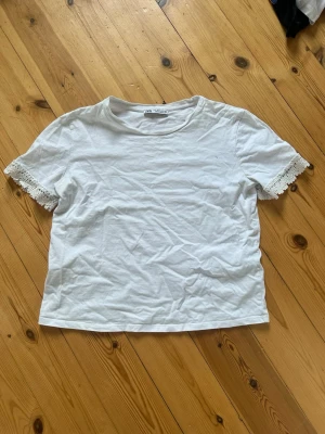 Vit t-shirt med spetsdetalj Zara - Säljer en vit t-shirt från Zara med korta ärmar och snygga spetsdetaljer vid ärmsluten. T-shirten har en klassisk rund halsringning och är gjord i mjuk bomull. Perfekt för dig som gillar enkla plagg med en twist!
