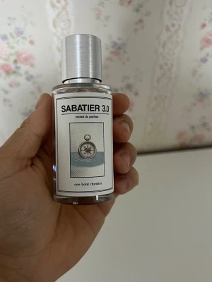 Sabatier 3.0 Extrait de Parfum - Säljer en stilren parfymflaska från Sabatier, modell 3.0. Flaskan är genomskinlig med en silverfärgad metallkork och sprayfunktion. Etiketten har en kompassillustration och texten 'one bold chemist'. Perfekt för dig som gillar unika dofter.