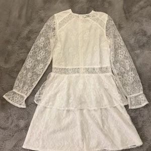 Vit spetsklänning från NA-KD studentklänning  - Säljer en elegant vit spetsklänning från NA-KD i storlek 34. Klänningen har långa spetsärmar och volangdetaljer i kjolen. Perfekt för speciella tillfällen, som till exempel studenten, med sin romantiska design.