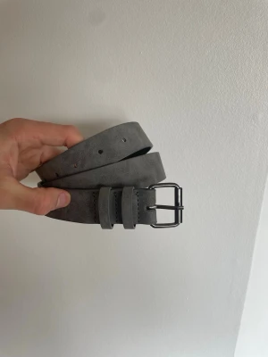Grått mockabälte med metallspänne - Snyggt grått bälte i mocka med klassiskt metallspänne. Bältet har en stilren design med dubbla hällor och justerbara hål. Perfekt accessoar för att lyfta din outfit.