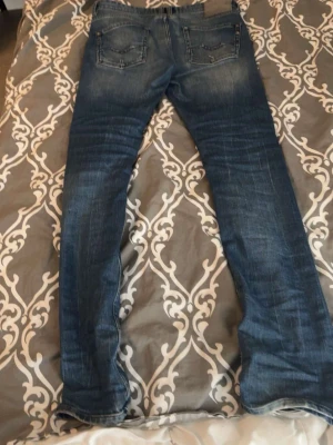 Blå raka jeans med slitningar - Säljer ett par klassiska blå jeans med raka ben och snygga slitningar. Jeansen har två bakfickor med dekorativa sömmar och en ljus tvätt för en avslappnad look. Perfekta till vardags och passar till det mesta. Materialet är troligtvis bomull med stretch.