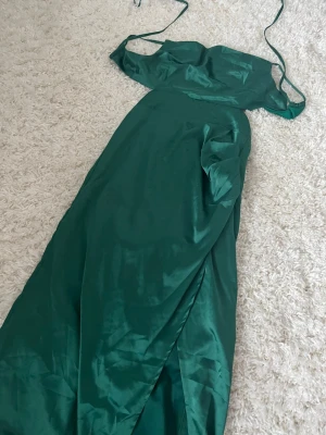 Grön långklänning i satin - Elegant grön långklänning i glansigt satinmaterial med axelbandslös design och smala knytband. Klänningen har en slits framtill och en figurnära passform som ger en snygg siluett. Perfekt för speciella tillfällen.