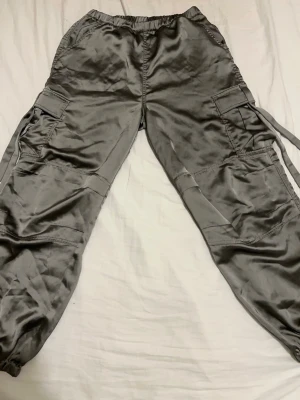 Grön/grå/ silvriga cargobyxor med fickor - Säljer ett par grön/grå cargobyxor med bred resår i midjan och flera stora fickor på sidorna och baktill. Byxorna har avslappnad passform och snördetaljer på benen. Perfekta för en streetwear-look och riktigt sköna att bära.