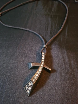 Svart korshalsband med strass från Z&R - Snyggt svart halsband med korsformad hängare dekorerad med gnistrande strass. Hängaren har en modern, böjd design och halsbandet är av mörk snodd. Perfekt för dig som gillar stilrena accessoarer med lite edge. Band knyts för önskad längd.