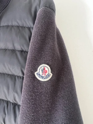 Svart cardiganjacka från Moncler - Snygg svart cardiganjacka från Moncler med stickade ärmar och vadderad framsida. Jackan har Moncler-logga på ärmen och ribbade muddar. Perfekt för dig som vill ha en exklusiv och trendig look.