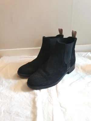 Svarta Chelsea boots i mocka - Snygga svarta Chelsea boots i mocka med klassisk siluett och elastiska paneler på sidorna. Bootsens dragflikar bak är i kontrasterande brunt läder för en cool detalj. Perfekta för dig som vill ha stilrena och tidlösa skor till höst och vinter.