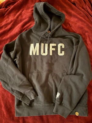 Svart MUFC hoodie Manchester United - Säljer en svart hoodie med stor MUFC-text på bröstet och Manchester Uniteds klubbmärke på ärmen. Hoodien har huva med dragsko och en stor magficka. Perfekt för dig som vill representera ditt favoritlag på ett stilrent sätt.