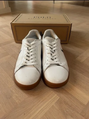 Vita Polo Ralph Lauren sneakers - Snygga vita sneakers från Polo Ralph Lauren med bruna gummisulor och mörkblå detaljer vid hälen. Klassisk låg modell med snörning och diskret logga på sidan. Perfekta för en stilren och sportig look. Använd ett par gånger. Bra skick bara några defekter men nästan som nya. Storlek 41 pris kan diskuteras