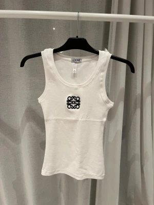 Vit linne från Loewe - Snygg vit linne från Loewe med broderad svart logga framtill. Toppen är ärmlös och har en klassisk rund halsringning. Perfekt för en stilren och trendig look. Tillverkad i mjuk bomull för maximal komfort.