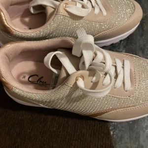 Glittriga beige sneakers från Clou - Snygga sneakers från Clou i beige med glittrig ovandel och vita snören. Skorna har detaljer i ljusrosa och guld, samt en vit sula. Perfekta för dig som vill sticka ut med lite extra bling i vardagen.
