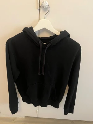 Svart hoodie från H&M, storlek S - En klassisk svart hoodie från H&M i storlek S. Hoodien har huva med dragsko, magficka och ribbade muddar vid ärmslut och nederkant. Perfekt för en avslappnad och stilren look. Tillverkad i mjukt material för extra komfort.