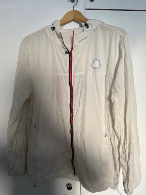 Vit vindjacka från Moncler - Snygg vit vindjacka från Moncler med huva och dragsko. Jackan har en röd och marinblå dragkedja framtill samt två fickor med dragkedja. Moncler-logga på bröstet. Tillverkad i lätt och tunt material, perfekt för blåsiga dagar.   