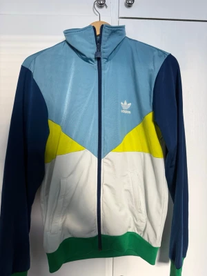 Adidas färgglad ziptröja - Snygg ziptröja från Adidas med färgblock i ljusblått, mörkblått, vitt, gult och grönt. Tröjan har hög krage, dragkedja framtill och två fickor. Adidas-logga på bröstet. Perfekt för en sportig och trendig look. Retro Tröja, 