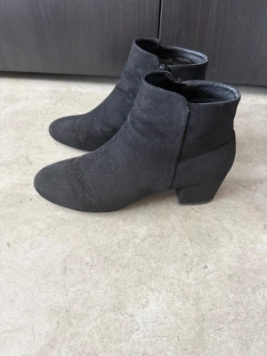 Svarta mockastövletter med klack - Snygga svarta stövletter i mockaimitation med rund tå och bred klack. Dragkedja på insidan för enkel på- och avtagning. Perfekta till jeans eller kjol för en trendig look.