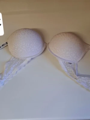 Vit push-up bh med spetsdetaljer - Säljer en vit push-up bh med vadderade kupor och fina spetsdetaljer längs banden. Bh:n har ett diskret mönster på kuporna och breda spetsband som ger en romantisk känsla. Perfekt för dig som vill ha både komfort och stil.