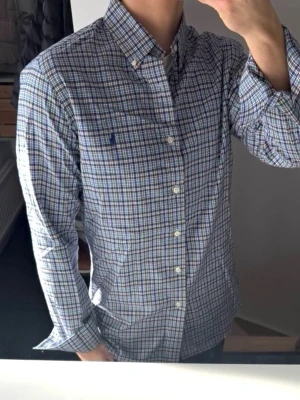 Rutig skjorta från Polo Ralph Lauren - Säljer en klassisk långärmad skjorta från Polo Ralph Lauren med rutigt mönster i blått, grönt och vitt. Skjortan har knappar framtill, button-down krage och en broderad logga på bröstet. Perfekt för dig som gillar stilrena och tidlösa plagg.