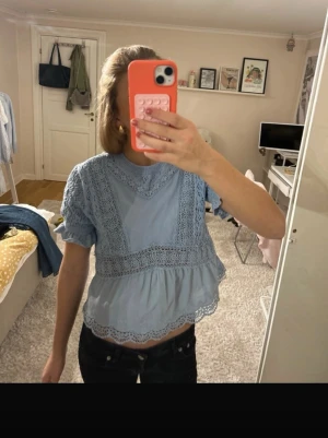 Ljusblå spetsblus med volang - Supersöt ljusblå blus med spetsdetaljer och volangkant. Blusen har korta ärmar med volang och rund halsringning. Perfekt för en trendig och feminin look. Materialet känns lätt och luftigt.