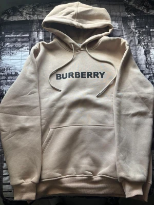 Beige hoodie från Burberry - Snygg beige hoodie från Burberry med klassisk logga tryckt på bröstet. Tröjan har huva med dragsko, magficka och ribbade muddar vid ärmslut och nederkant. Perfekt för en avslappnad och trendig stil.