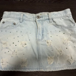 Ljus jeanskjol med nitar och slitningar - Snygg ljusblå jeanskjol med slitna detaljer och glittrande nitar framtill och på bakfickorna.Midjemåttet rakt över är 37 cm. Obs! Jag är på semester och kan därmed skicka tidigast 28/6💓