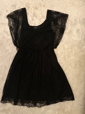 Svart spetsklänning med korta fjärilsarmar och vidd nedtill - Snygg svart kortklänning med transparenta spetsärmar och rund halsringning. Klänningen har en lätt utställd kjol och är tillverkad i ett tunt, mjukt material med spetsdetaljer som ger en romantisk vibe. Perfekt för dig som gillar en feminin och trendig stil. Längd 87 cm.