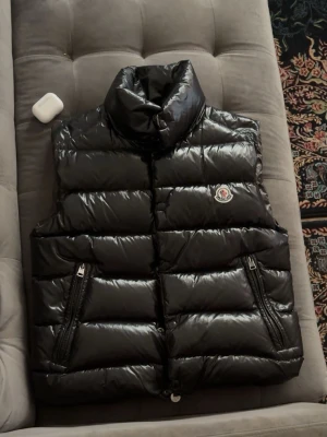 Svart dunväst från Moncler - Snygg svart dunväst från Moncler med glansig finish och två dragkedjefickor framtill. Klassisk pufferstil med Moncler-logga på bröstet och hög krage. Perfekt för lager-på-lager och streetwear.