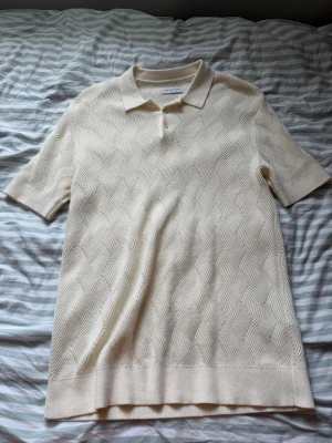Beige stickad polotopp från Arket - Snygg beige stickad polotopp från Arket med korta ärmar och klassisk krage. Toppen har ett diskret vågigt mönster i materialet och är perfekt för en clean och stilren look. Jätte fint skick och lite finare tröja men kan diskutera pris:)  storlek runt M