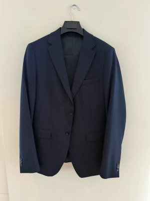 Mörkblå kostym med kavaj och byxor - Snygg mörkblå kostym med klassisk kavaj och matchande byxor. Kavajen har två knappar framtill, fickor med lock och smal krage. Perfekt för dig som vill ha en stilren och tidlös look.