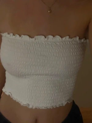 Vit smockad bandeau tubtopp - Säljer en vit bandeau tubtopp med smockad struktur och volangkant upptill och nedtill. Toppen är axelbandslös och har en tight passform som sitter snyggt på kroppen. Perfekt för varma dagar eller som layering-piece.
