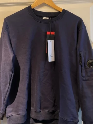 Mörkblå sweatshirt från C.P. Company - Säljer en mörkblå sweatshirt från C.P. Company. Tröjan har den ikoniska linsen på ärmen. Perfekt för dig som gillar streetwear och stilrena detaljer. Tröjan är aldrig använd och säljes för att den ej passar. Priset kan diskuteras vid en snabb affär🤝
