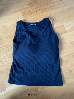 Svart ribbad linnetopp - En svart ribbad linnetopp med rund halsringning. Toppen är ärmlös och har en enkel, stilren design som passar perfekt till jeans eller kjol. Materialet känns mjukt och stretchigt, vilket gör den bekväm att bära hela dagen. Har en inbyggd bh.