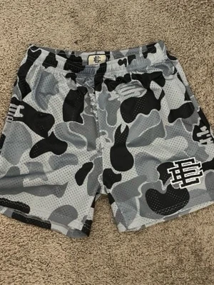 Grå camo shorts från EE - Snygga shorts i grått och svart camouflagemönster från EE. De har elastisk midja och är tillverkade i ett ventilerande meshmaterial. Perfekta för en avslappnad streetwear-look.