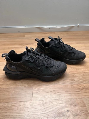 Svarta Nike React Vision - Säljer ett par helsvarta Nike React Vision sneakers med mesh- och syntetmaterial, chunky sula och genomskinliga detaljer. Skorna har en sportig look med dragflik bak och perforerat material för extra ventilation. Perfekta för dig som gillar streetwear och modern design.