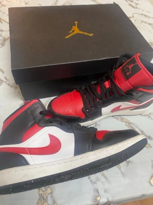 Air Jordan 1 Mid  - Snygga Air Jordan 1 Mid sneakers i rött, svart och vitt läder med klassisk Nike Swoosh på sidan och Air Jordan-logga vid ankeln. Skorna har svart snörning, vadderad plös och hög siluett som ger en ikonisk streetstyle-look. Jätte fina skor. Är typ helt nya. Orginal låda följer med 