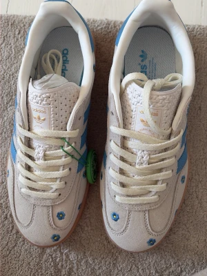 Adidas gazelle sneakers med blå blommor - Unika Adidas, gazelle sneakers i beige mocka med blå detaljer och små broderade blommor. Skorna har klassiska blå ränder på sidorna, vit plös med Adidas-logga i guld och beige snörning. Yttersulan är i gummi och ger en retrovibe. Perfekta för dig som vill sticka ut med din stil. Oanvända!! Beige/vita.