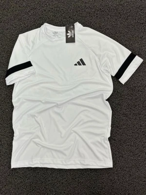 Vit Adidas topp med svarta detaljer - Vit topp från Adidas med korta ärmar och svarta ränder på ärmarna. Tillverkad i ett lätt och syntetiskt material, perfekt för en sportig look. Adidas-logga på bröstet och klassisk rund hals.